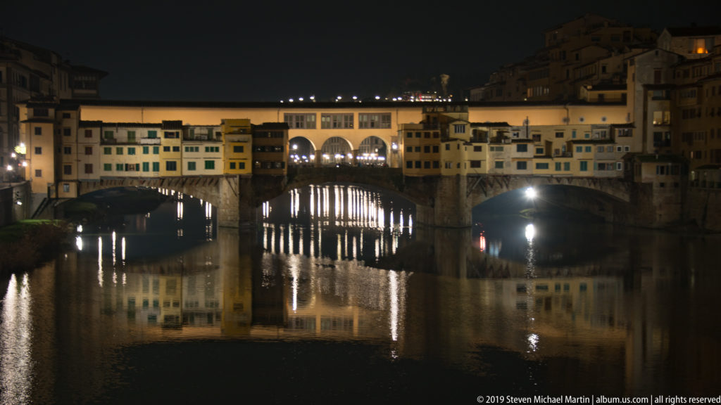 Close nighttime view of Ponte Vecchio with illuminated buildings above stone arches reflected in the Arno River.