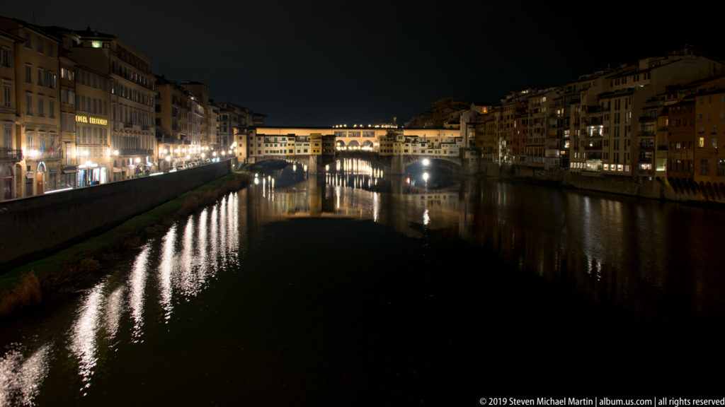 Night view along the Arno toward illuminated Ponte Vecchio, with riverfront buildings and lights reflected in the water.
