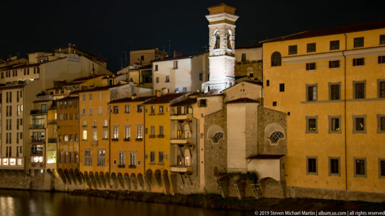 Illuminated ochre riverfront buildings and a small church with bell tower beside the dark Arno River at night.