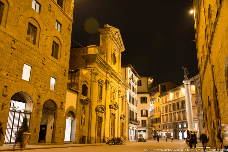 Oblique night view of a historic Florence church beside a pedestrian square lined with lit buildings and shops.