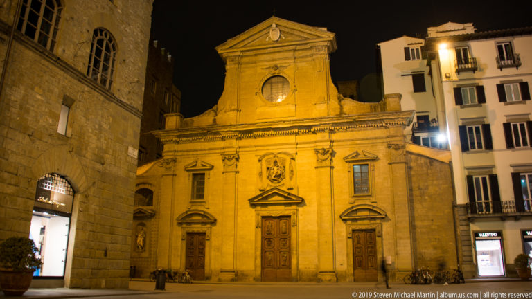 Amber-lit church façade with pilasters, triangular pediment, round window, carved emblems, and three doorways in Florence.