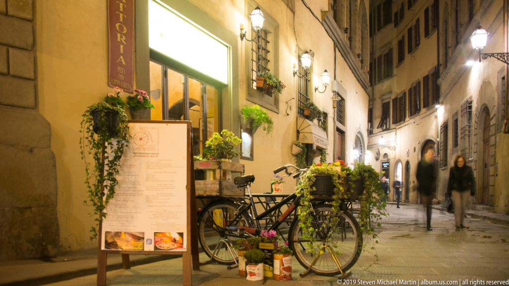 Narrow illuminated Florence street with restaurant menu, bicycles, potted plants, wall lamps, and pedestrians.
