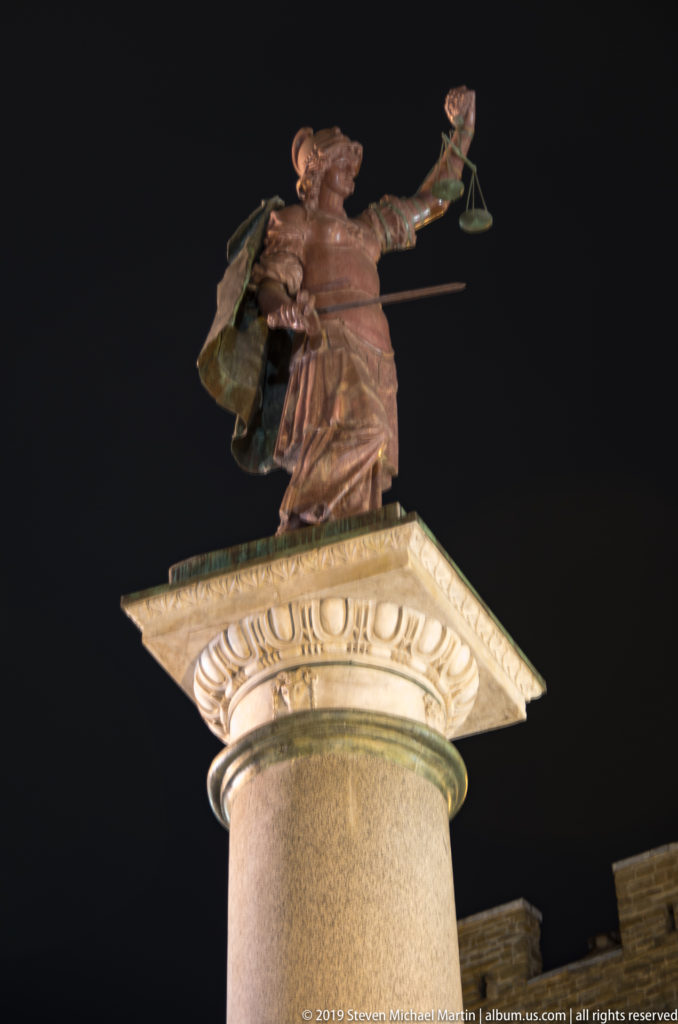 Bronze figure of Justice holding scales atop the tall Column of Justice against a black night sky.