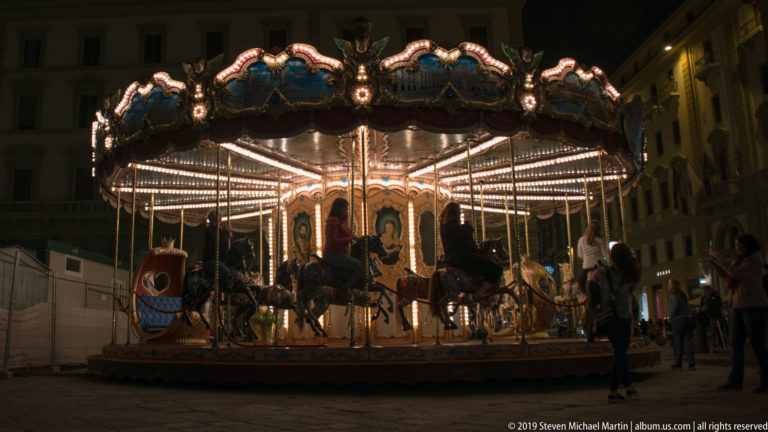 Brightly illuminated carousel with painted canopy and carved horses operating in a Florence square at night.