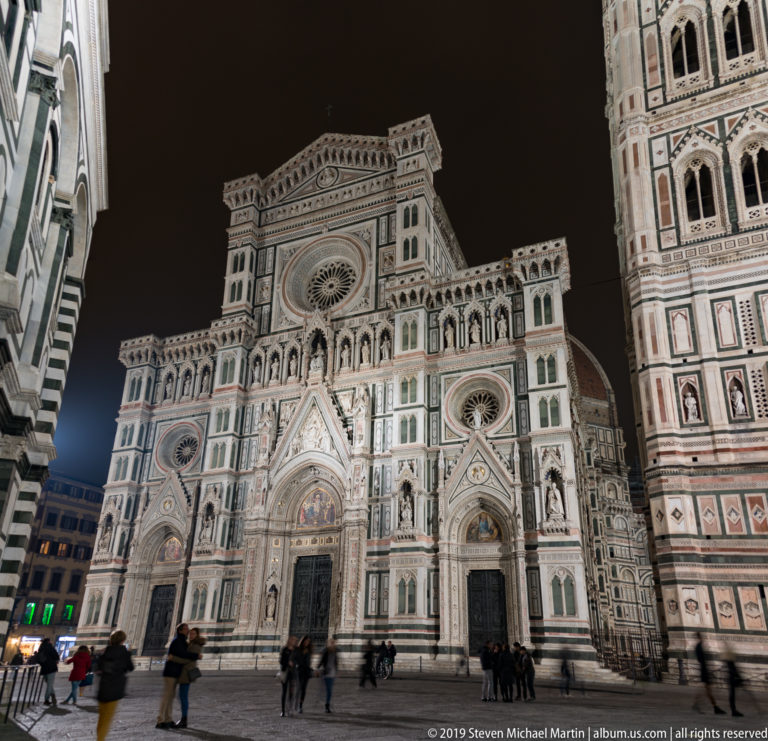 Nearly frontal nighttime view of Florence Cathedral across the piazza with pedestrians and part of Giotto’s Campanile.