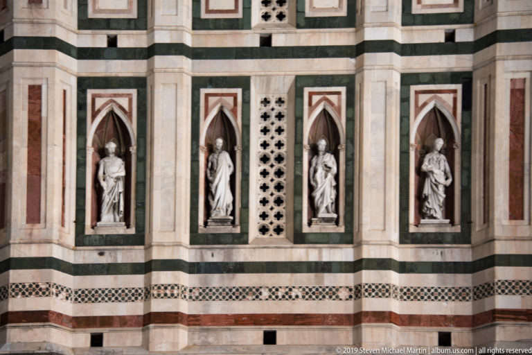 Three marble statues in pointed niches set into the polychrome lower façade of Giotto’s Campanile.