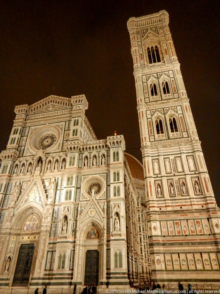 Illuminated Florence Cathedral façade beside the tall square Giotto’s Campanile against a dark night sky.