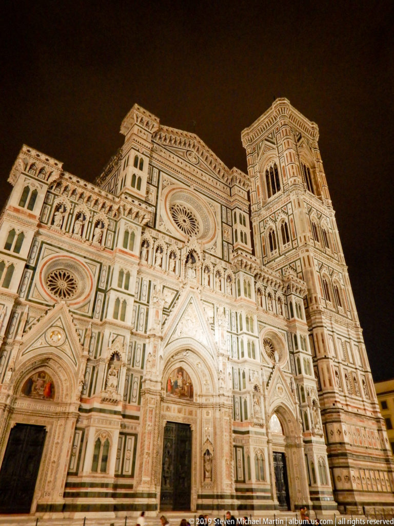 Full illuminated west façade of Florence Cathedral with three portals, rose windows, niches, and polychrome marble decoration.