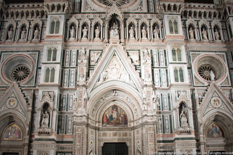 Central portal and surrounding sculptural, circular-window, and marble decoration on the illuminated façade of Florence Cathedral.