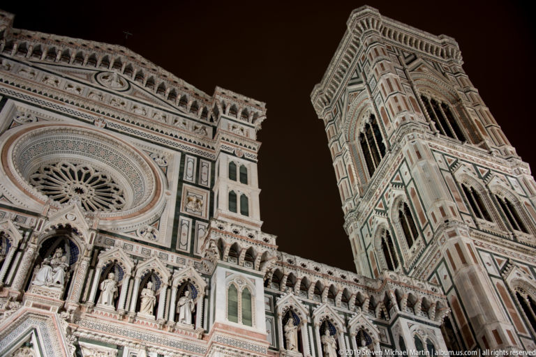 Illuminated marble façade of Florence Cathedral and Giotto’s Campanile seen from a steep upward angle at night.