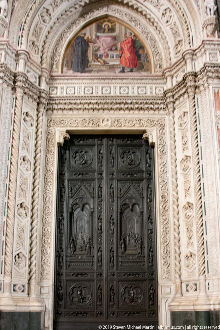 Full Florence Cathedral portal with dark bronze relief doors, mosaic lunette, and densely carved pale marble surround.