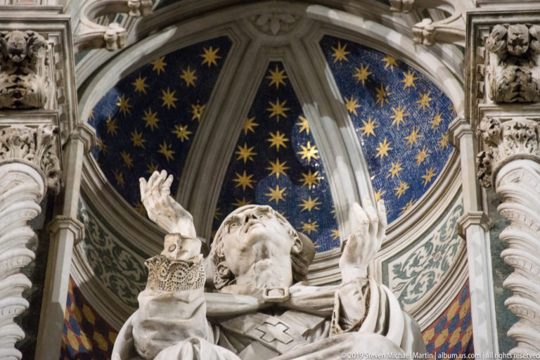 White marble figure raising both hands beneath a blue canopy painted with gold stars, framed by carved cathedral architecture.