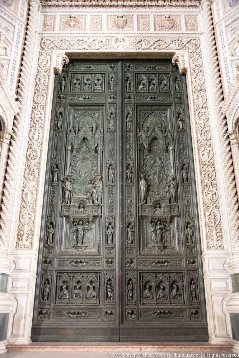 Pair of dark bronze relief doors centered within an ornate pale marble portal on Florence Cathedral.
