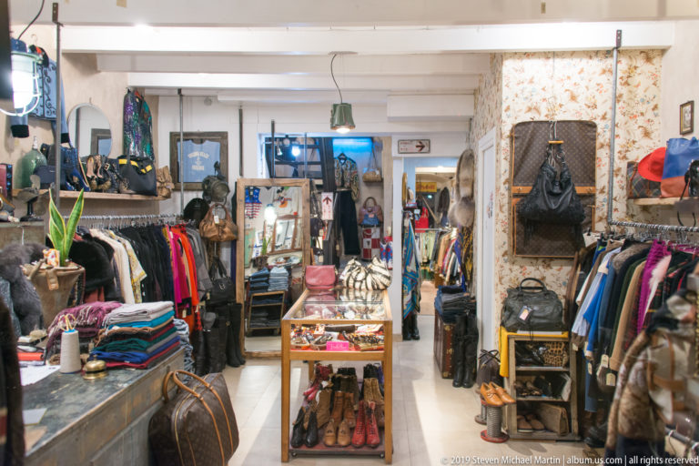 Narrow boutique packed with clothing racks, handbags, shoes, scarves, mirrors and display cases.