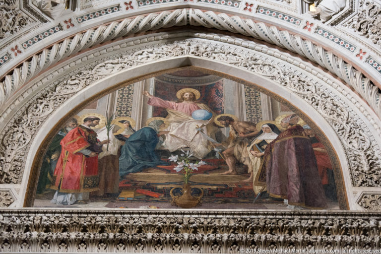 Enthroned haloed figure with attendants in a colorful lunette mosaic beneath densely carved pointed arches.