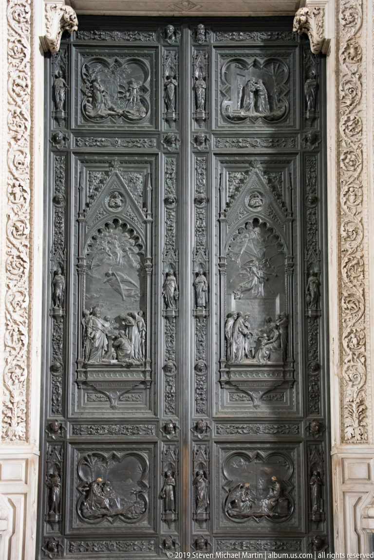Tall dark bronze cathedral door divided into sculpted relief panels, framed by pale carved marble ornament.