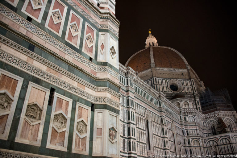 Oblique view along Florence Cathedral’s polychrome marble wall toward Brunelleschi’s illuminated dome and lantern at night.