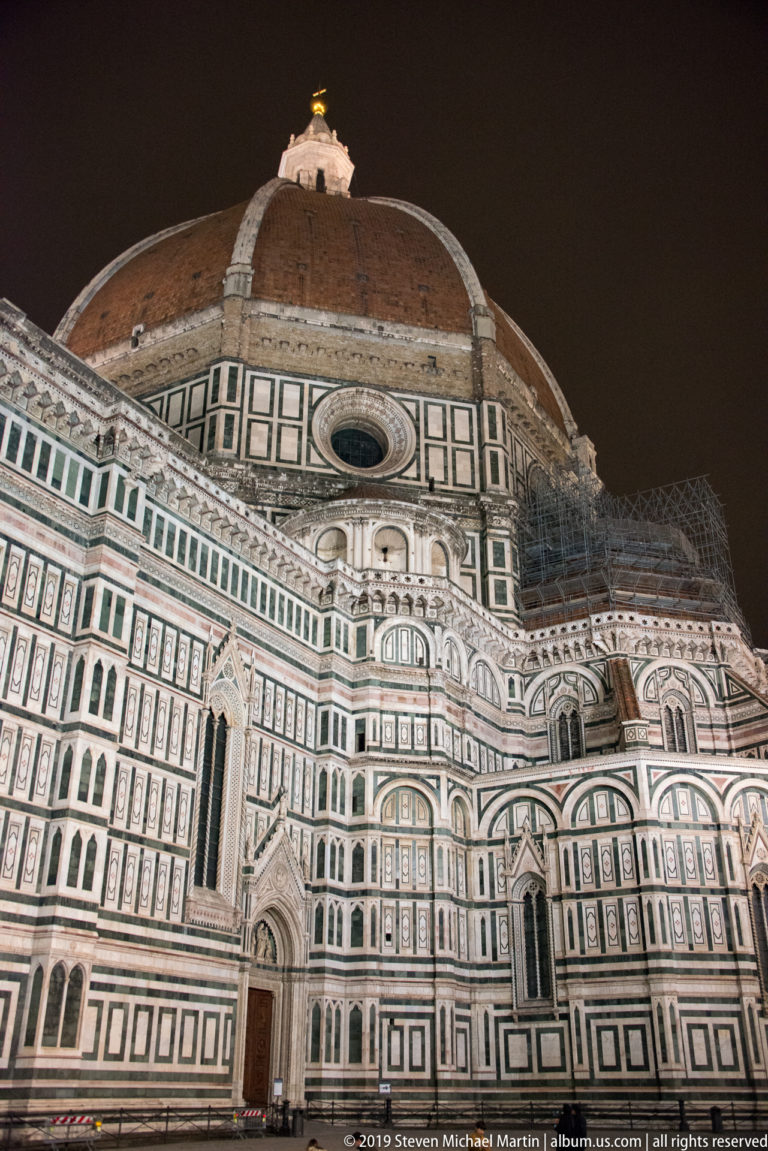 Night view along Florence Cathedral’s patterned marble side toward Brunelleschi’s large red-tiled dome, with scaffolding visible nearby.