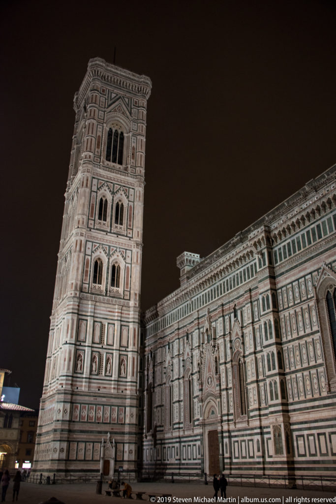 Illuminated Giotto’s Campanile beside the long polychrome marble side of Florence Cathedral at night.