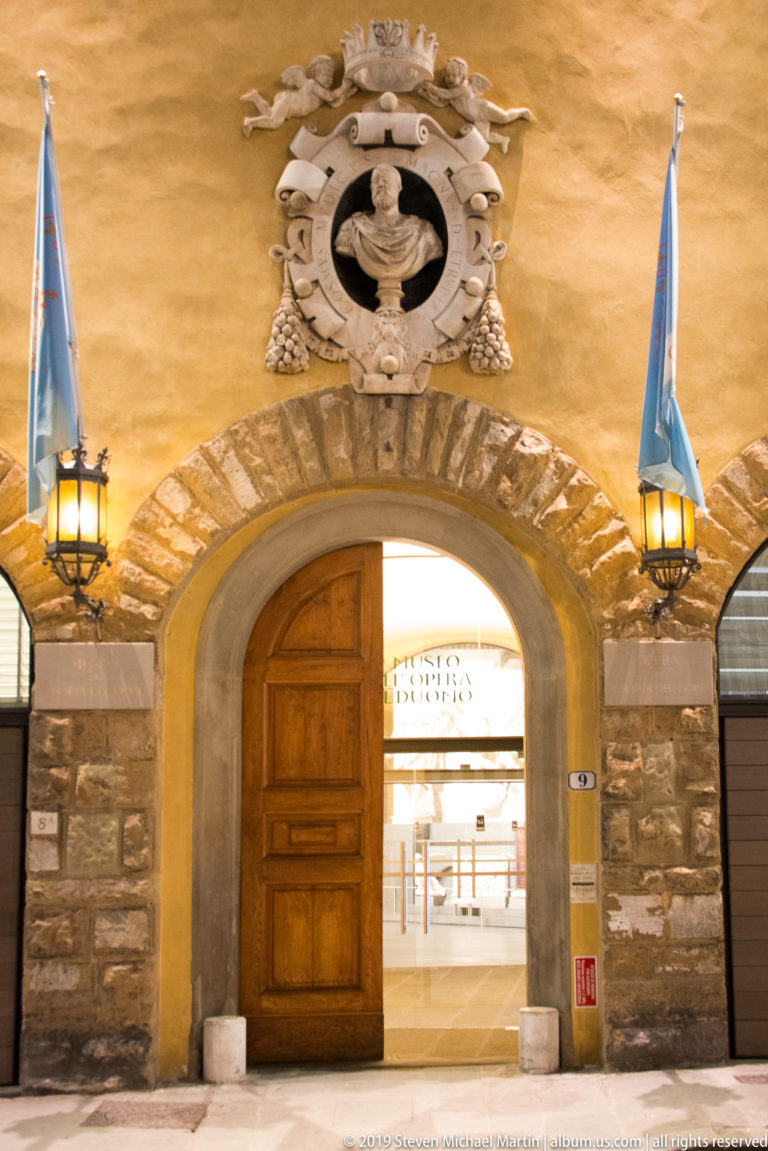Partly open wooden door beneath a carved heraldic crest, flanked by blue flags and lanterns on a warm ocher wall.