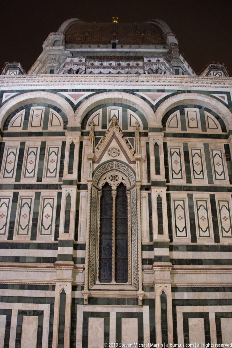 Tall Gothic window centered in Florence Cathedral’s green-and-white patterned marble wall under nighttime illumination.