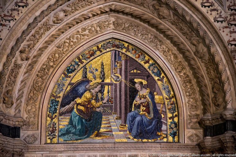 Annunciation mosaic with kneeling angel and seated Virgin beneath a pointed arch surrounded by densely carved marble ornament.