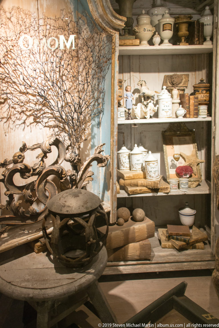 Branching metal tree sculpture before shelves of pale ceramics, bowls and decorative objects in a shop.