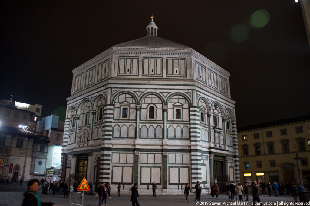 Illuminated octagonal Baptistery of San Giovanni with green-and-white marble panels and pedestrians in Piazza del Duomo at night.