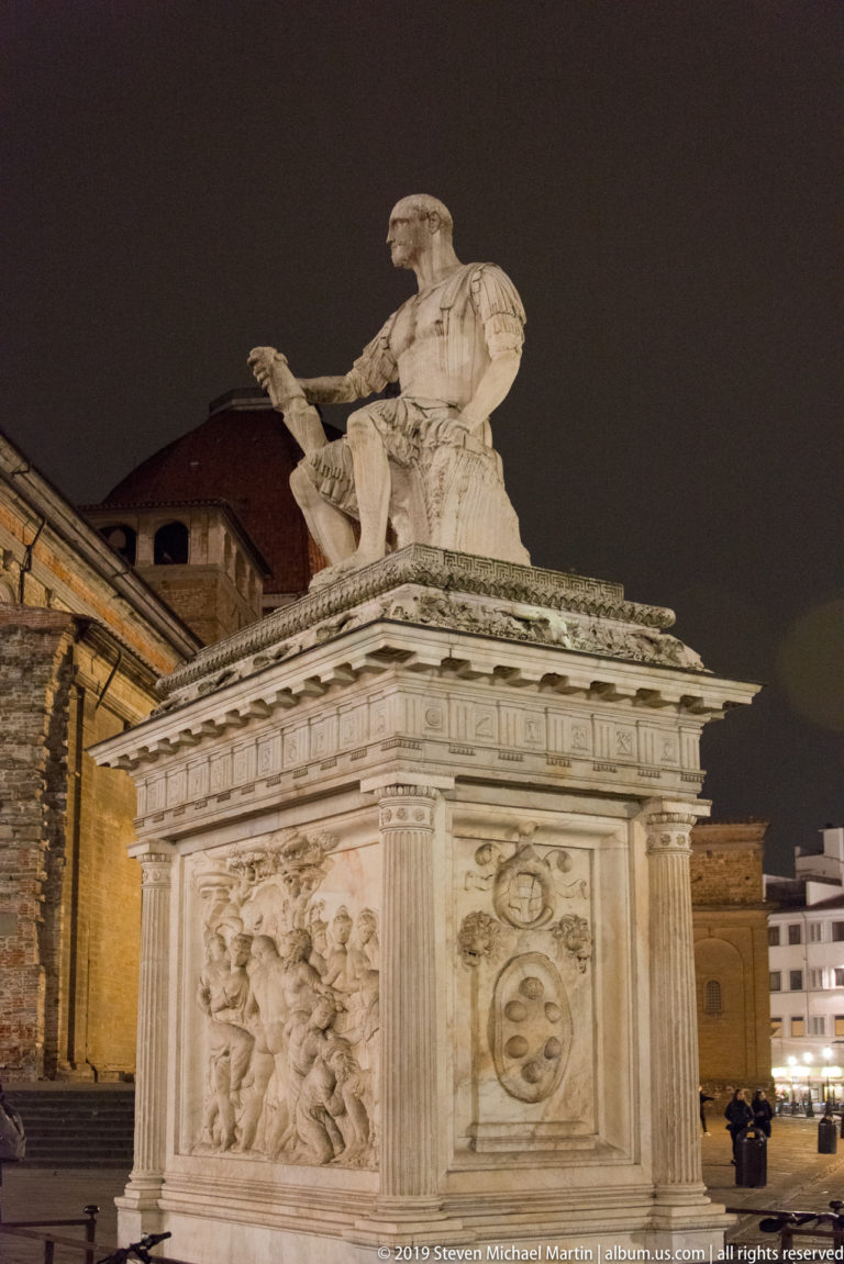 Night view of the seated armored figure of Giovanni delle Bande Nere above a carved stone pedestal with Medici emblems.