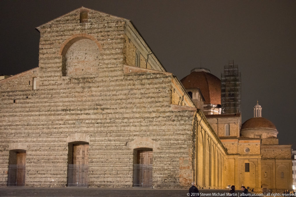Night view of San Lorenzo’s long rough-masonry side wall, tiled roof, domed structures, and scaffolding in Florence.