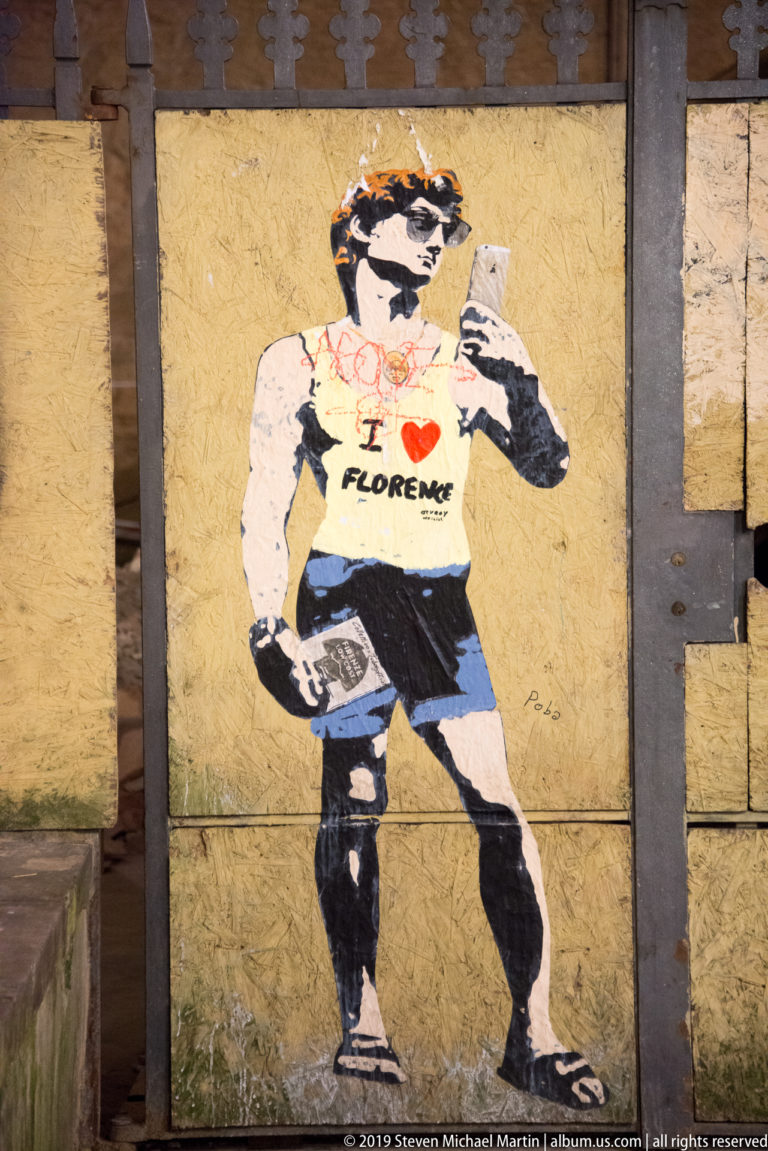 Stencil-style muscular man on an ocher wall wearing a white “I love Florence” shirt and dark briefs.