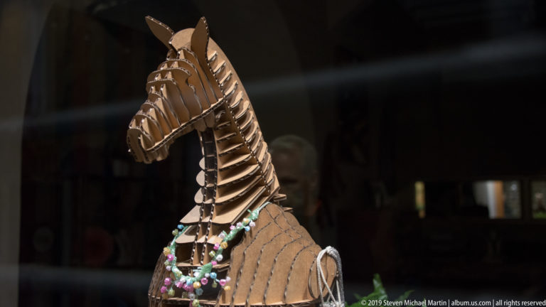 Openwork brown horse sculpture built from layered strips, with colorful bead strands hanging across its chest against a dark window.