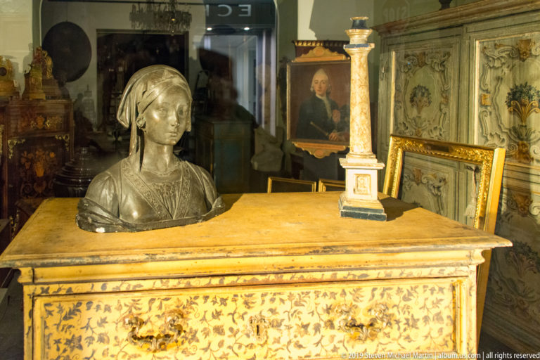 Pale female bust behind an inlaid wooden cabinet topped by a small column model, with framed art and antique furniture beyond.