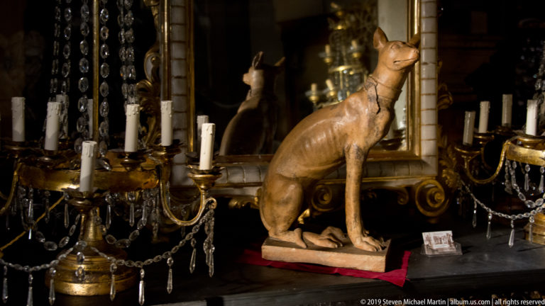 Seated brown cat sculpture among ornate candlesticks, crystal drops, and mirrors in a densely arranged interior display.