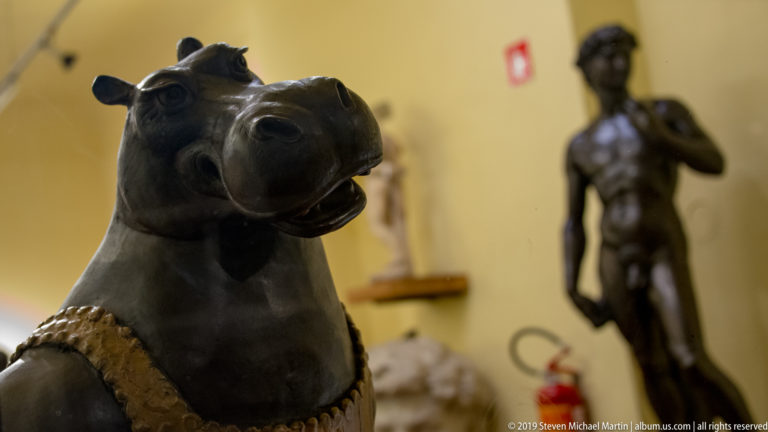 Close view of a dark bronze horse head with decorative tack, with a standing nude male sculpture blurred in the background.