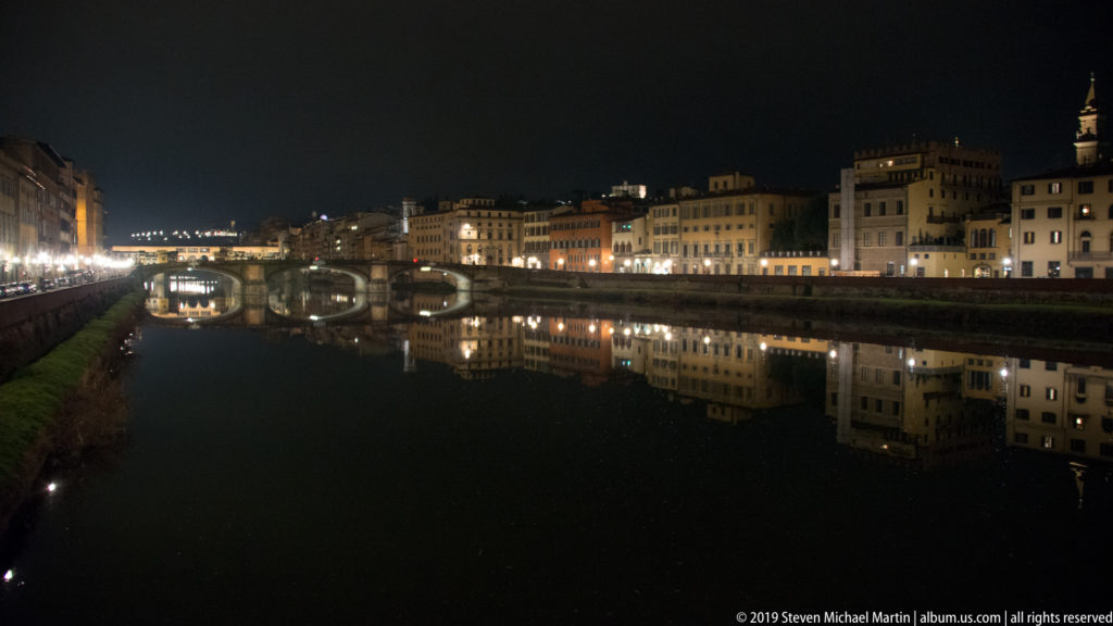 Night view along the Arno with illuminated bridges and historic riverfront buildings reflected in calm dark water.