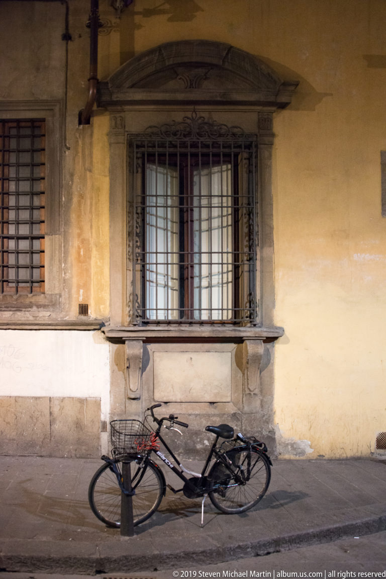 Bicycle leaning beneath a barred, stone-framed window in a weathered yellow plaster wall.