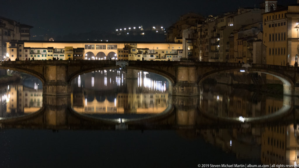 Illuminated Ponte Vecchio crossing the dark Arno, with shop-lined upper levels and bright reflections beneath its stone arches.