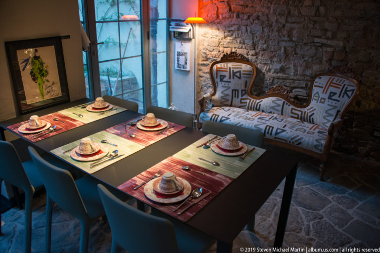 Table set for four with cups and placemats beside blue chairs, a patterned settee, exposed stone wall, and tall window.