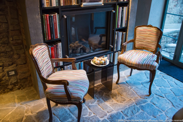 Two striped armchairs beside a small glass table, bookshelves, exposed stone walls, and a tall window in a warmly lit interior.