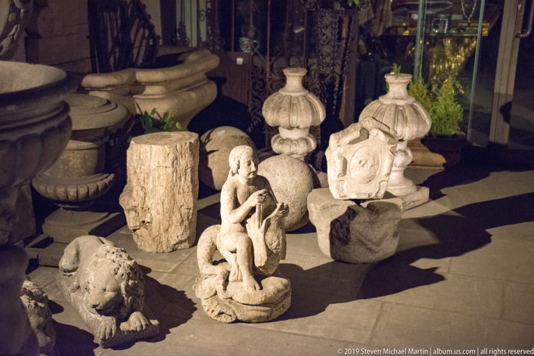 Carved stone garden ornaments including a seated child, animal figure, urns and finials under strong side lighting.
