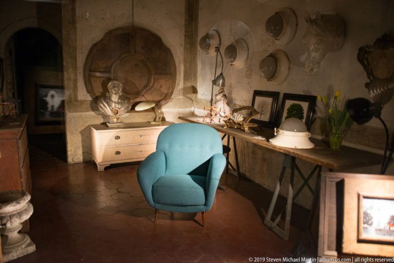 Turquoise armchair in an eclectic interior with wall-mounted objects, framed art, lamps and terracotta-colored floor tiles.