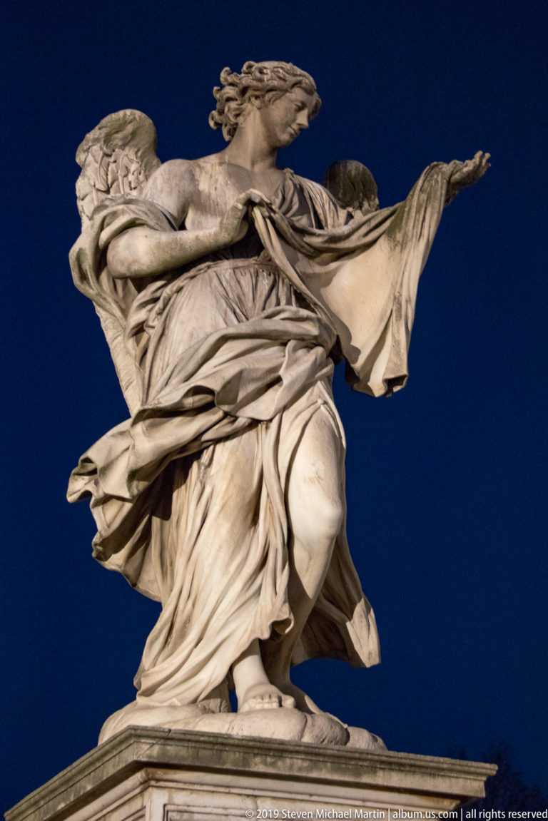 Floodlit marble angel on Ponte Sant’Angelo with one arm extended and wind-swept drapery against the dark sky.