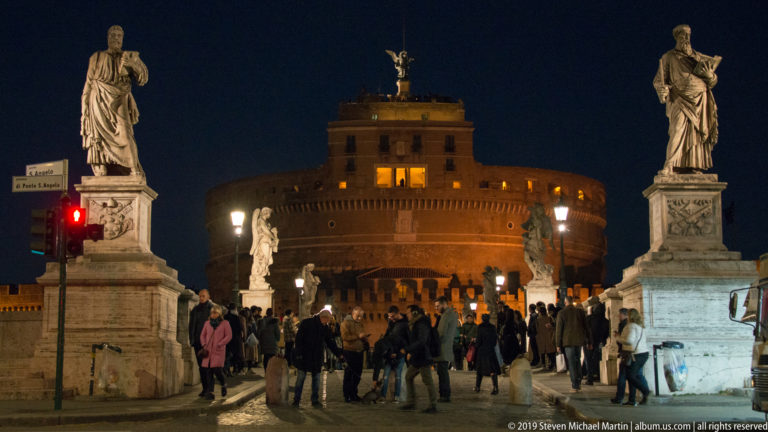 Castel Sant’Angelo illuminated beyond crowds and marble statues lining Ponte Sant’Angelo at night.