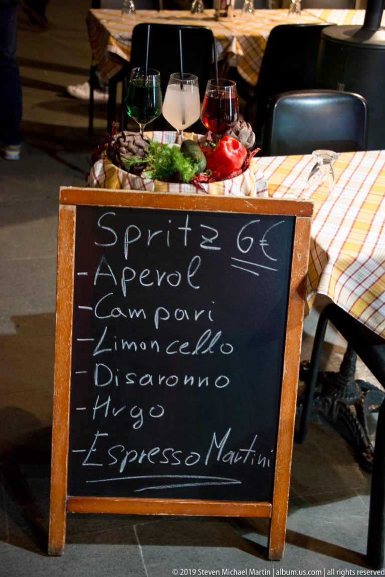 Chalkboard advertising six-euro spritz drinks beside an outdoor restaurant table set with glasses, plates, and food.