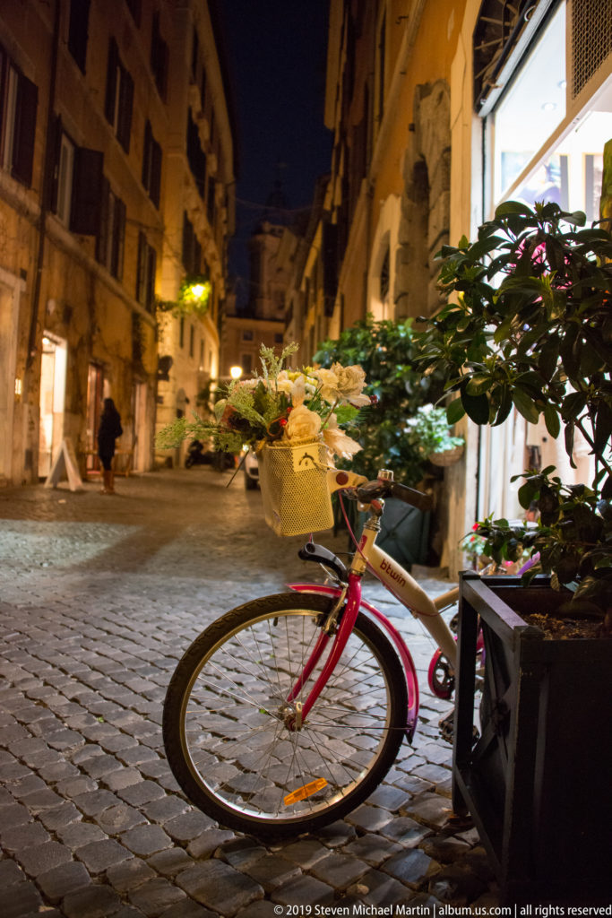 Red bicycle beside leafy planters and a bright shop on a narrow cobbled Roman street at night.
