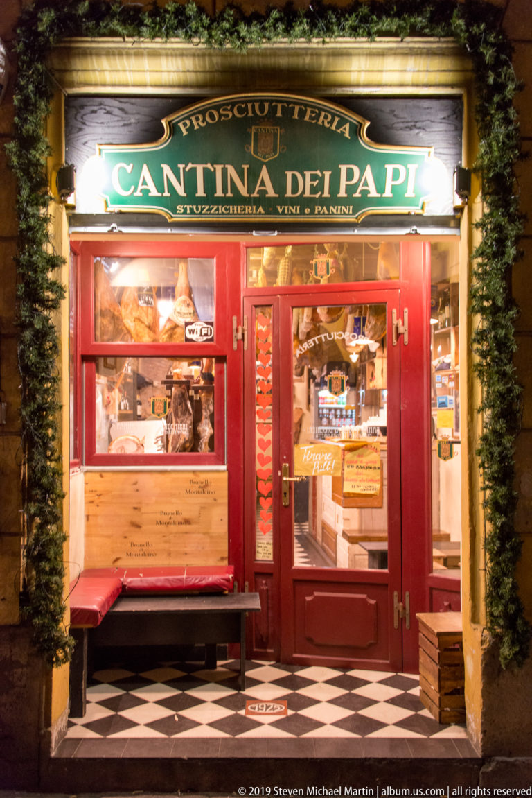 Bright red Cantina dei Papi prosciutteria storefront with illuminated signage, food displays, and black-and-white tiled entrance.
