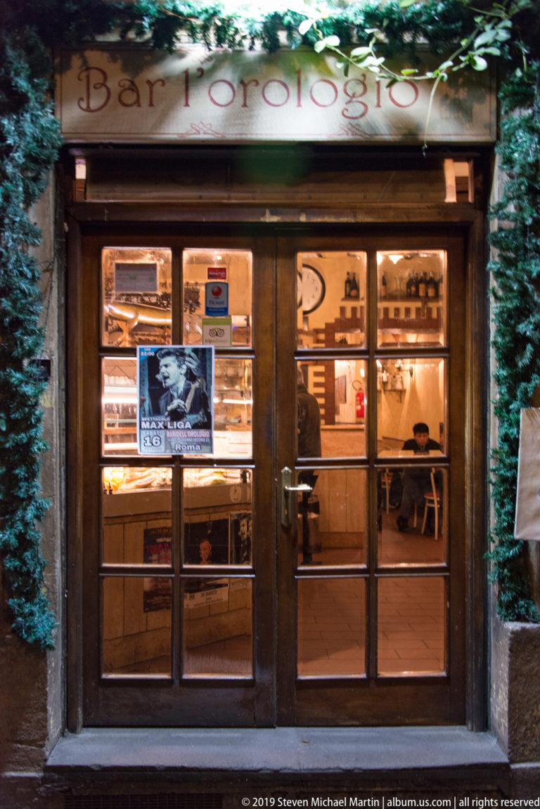 Wood-and-glass entrance to Orologio restaurant glowing warmly at night, framed by greenery with diners visible inside.