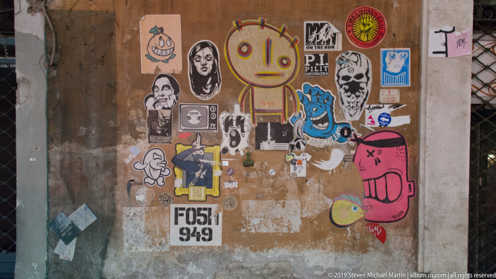 Weathered wall covered with layered stickers, cartoon figures, portraits, tags, and torn paper remnants.