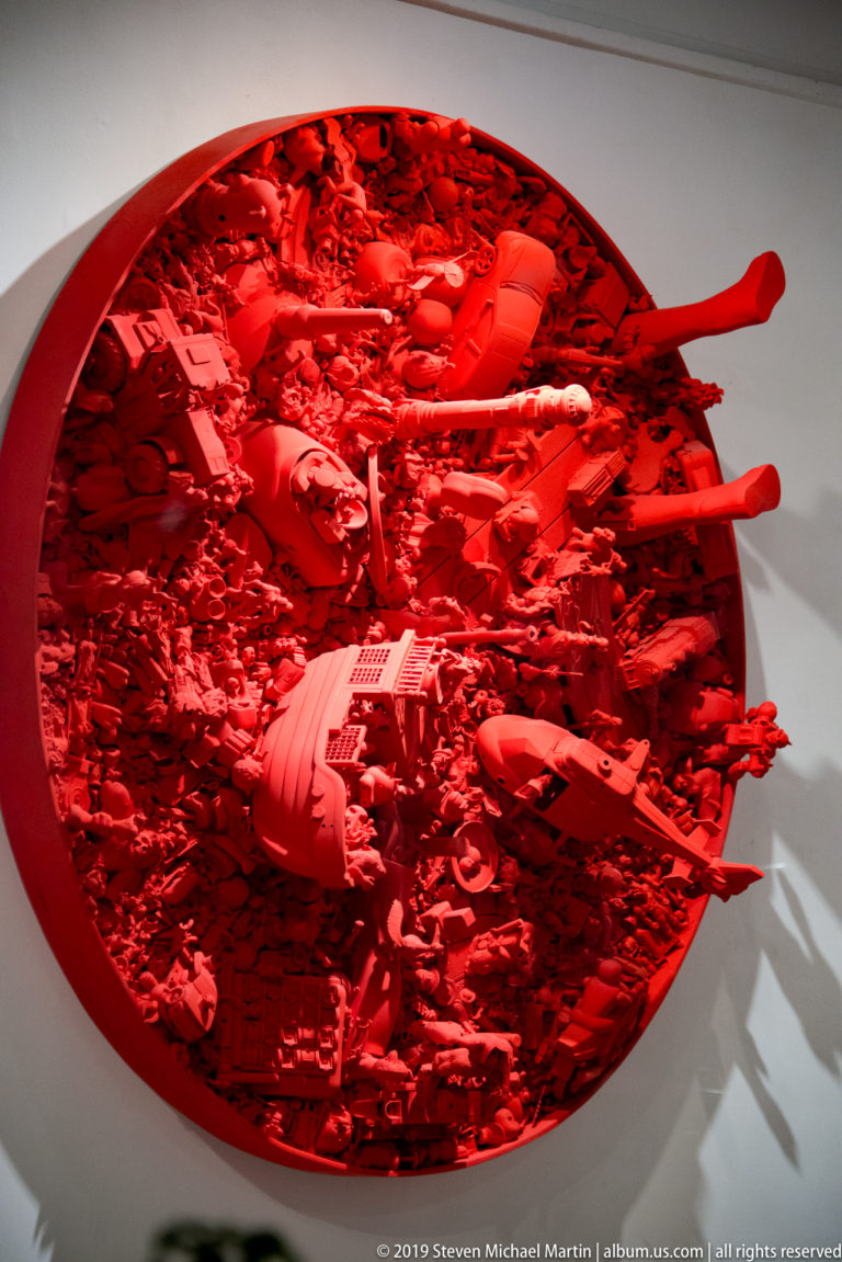Large circular red assemblage relief filled with embedded objects, including guitars and projecting paddles, mounted on a white wall.